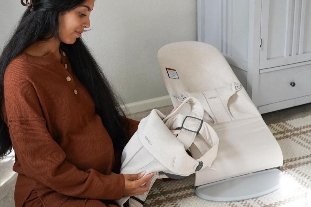 Pregnant woman in a brown sweater examines a beige baby bouncer.