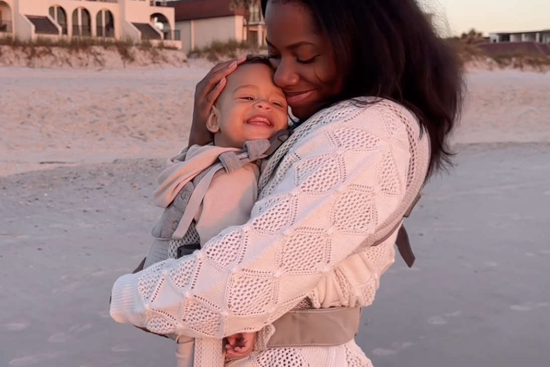 A smiling woman holds a happy baby in a carrier on a sandy beach at sunset, with buildings in the background.