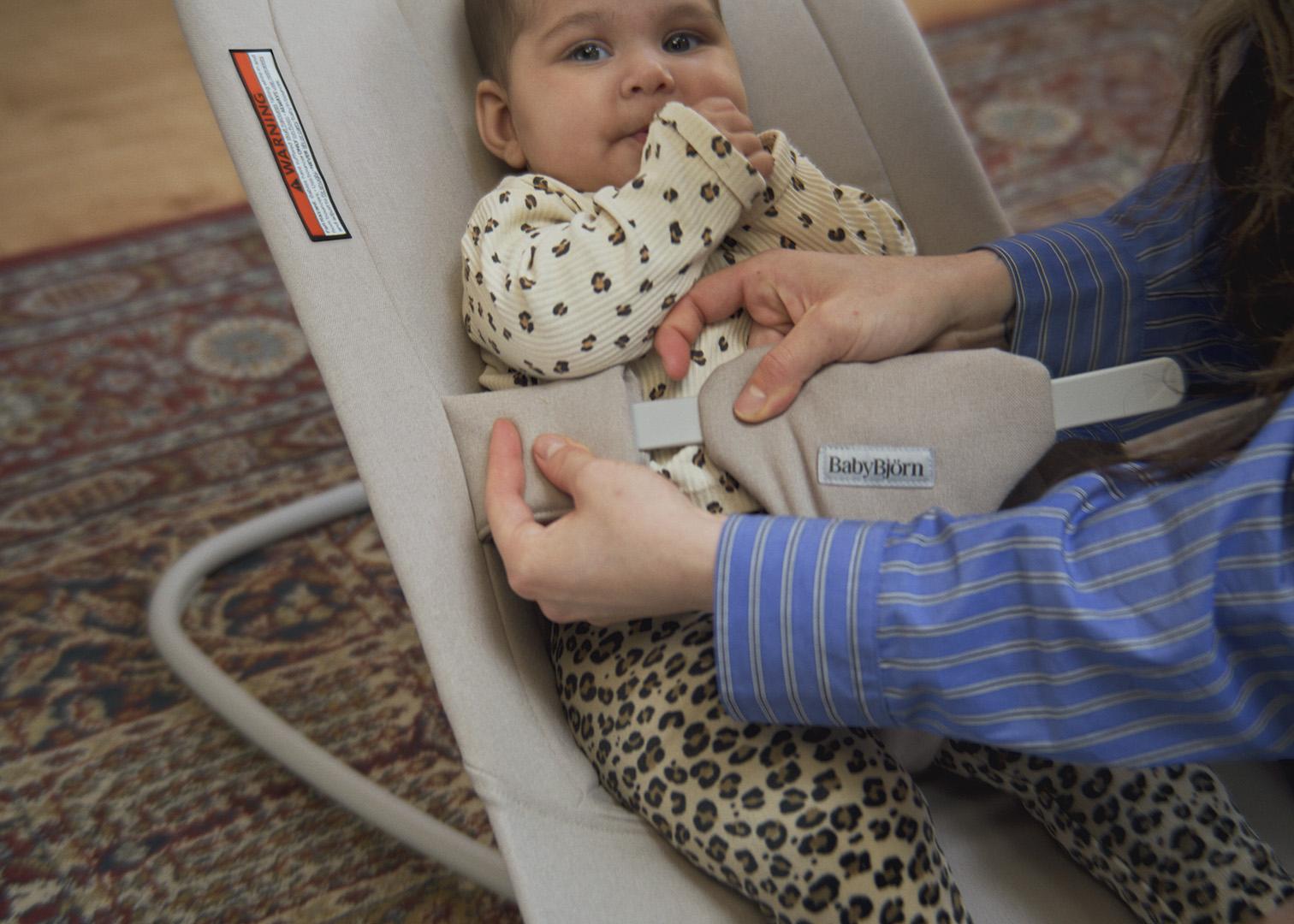 A baby is sitting in a beige bouncer with a parent securing the safety harness. The baby is wearing a patterned outfit on a patterned rug.
