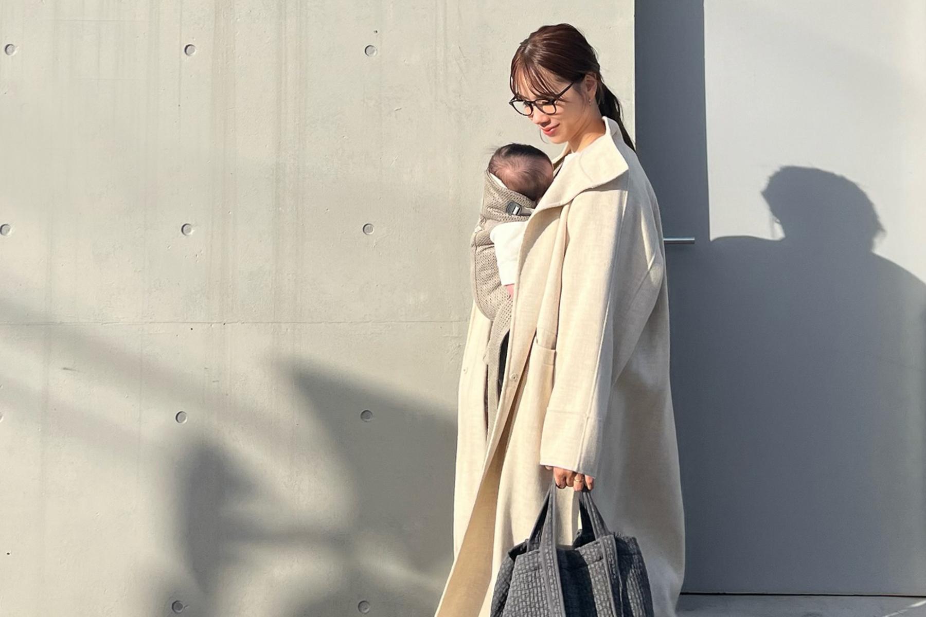 A person in a light coat carries a baby in a sling, holding a gray bag, standing against a minimalist concrete wall.