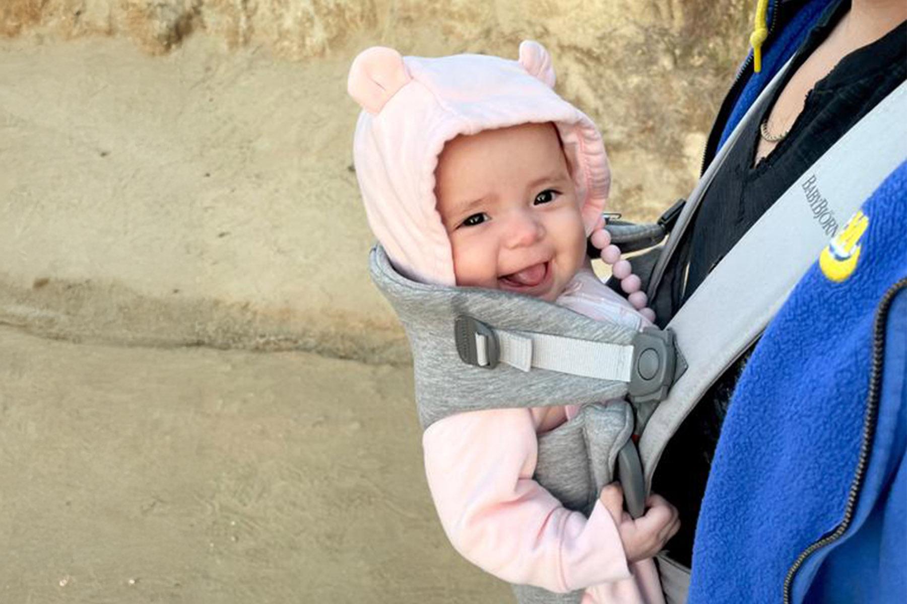 Smiling baby in a pink hooded outfit with bear ears in a gray front carrier, held by an adult wearing a blue jacket.