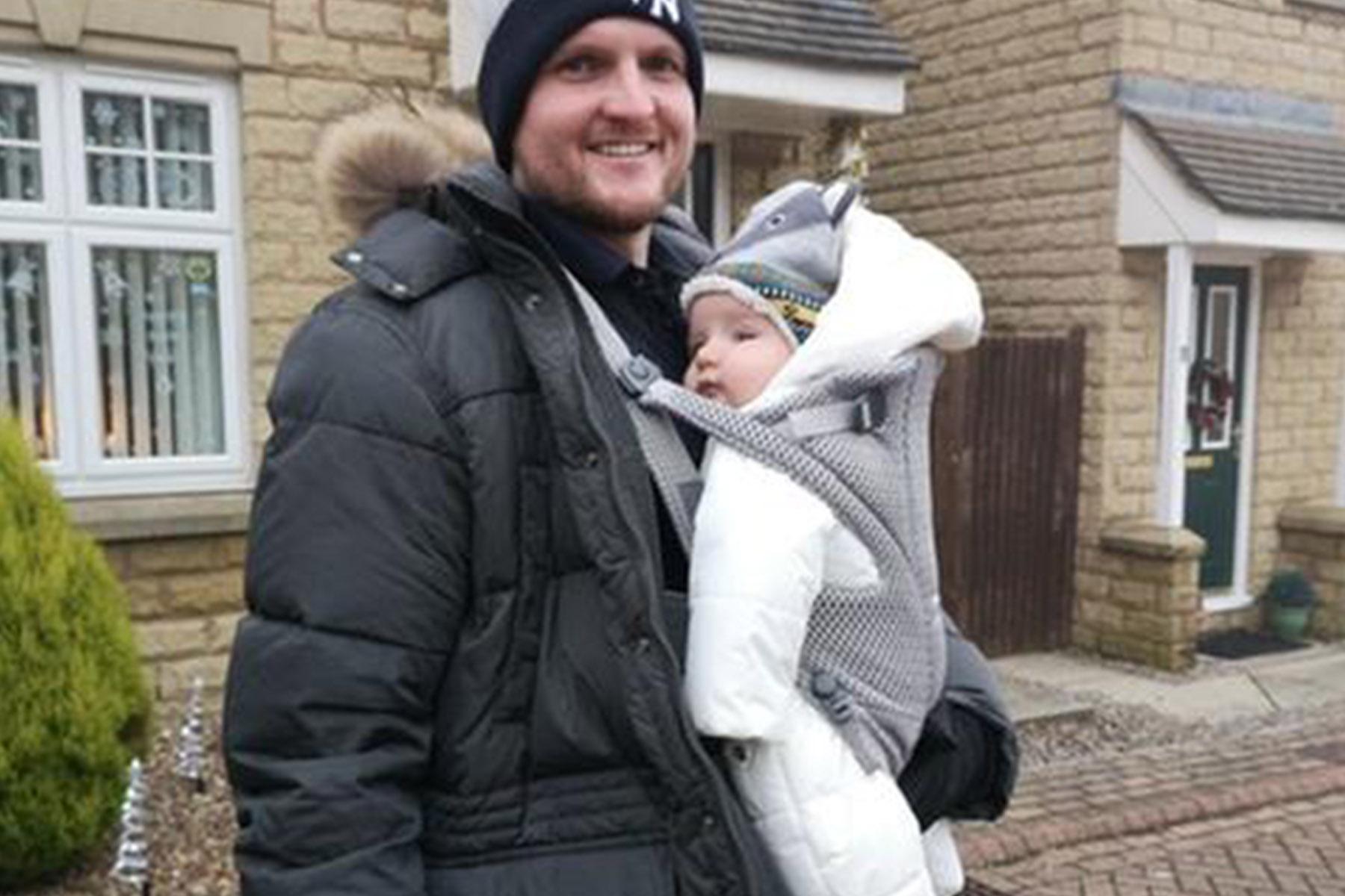 Man in winter coat and beanie smiling, carrying bundled baby in white snowsuit and knit hat in front of a stone house.