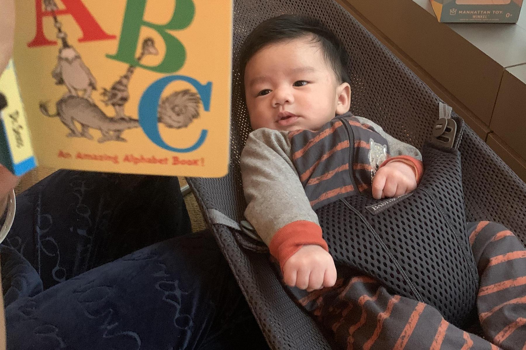 A baby in a striped outfit lies in a seat, looking at a colorful ABC book held by an adult.