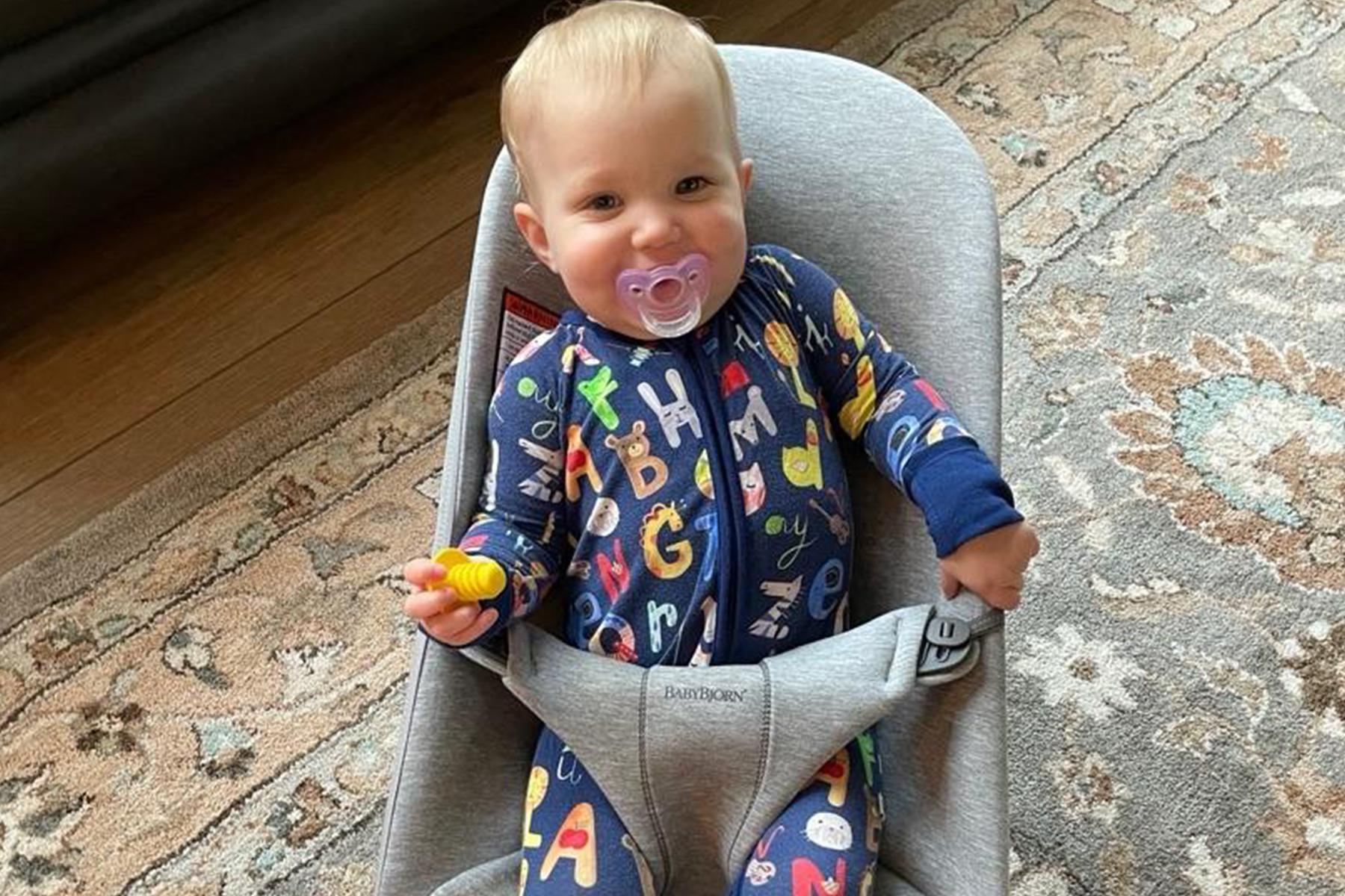 Baby in a blue alphabet-themed onesie sits in a gray baby bouncer, holding a yellow toy, with a pacifier in mouth, on a patterned rug.