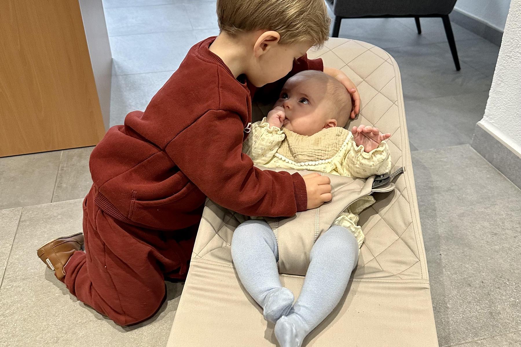 A young boy in a red outfit gently touches the head of a baby lying in a beige baby seat, showing affection.