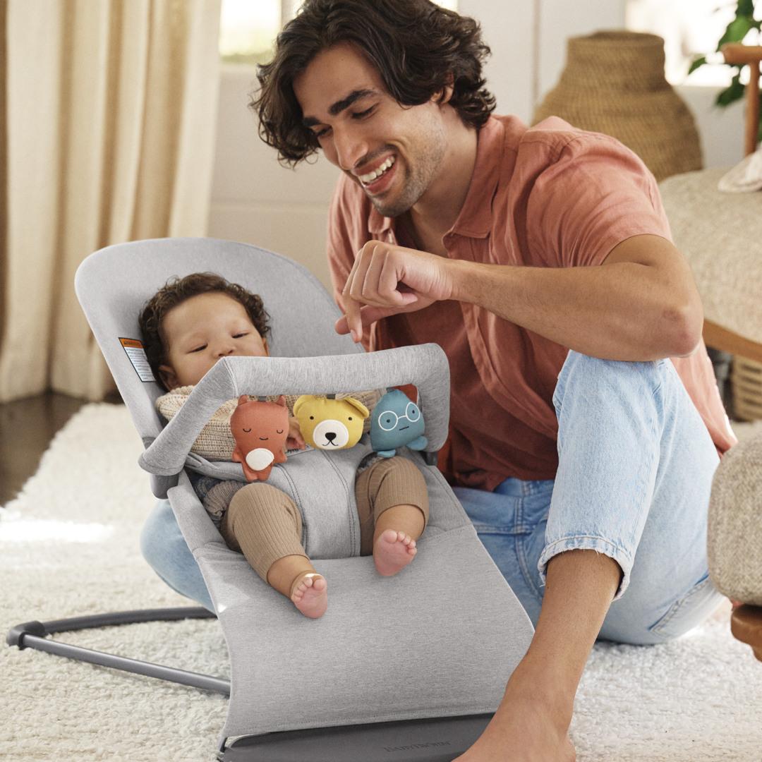 A smiling man interacts with a baby in a gray bouncer, surrounded by plush toys. The room is cozy with warm lighting.