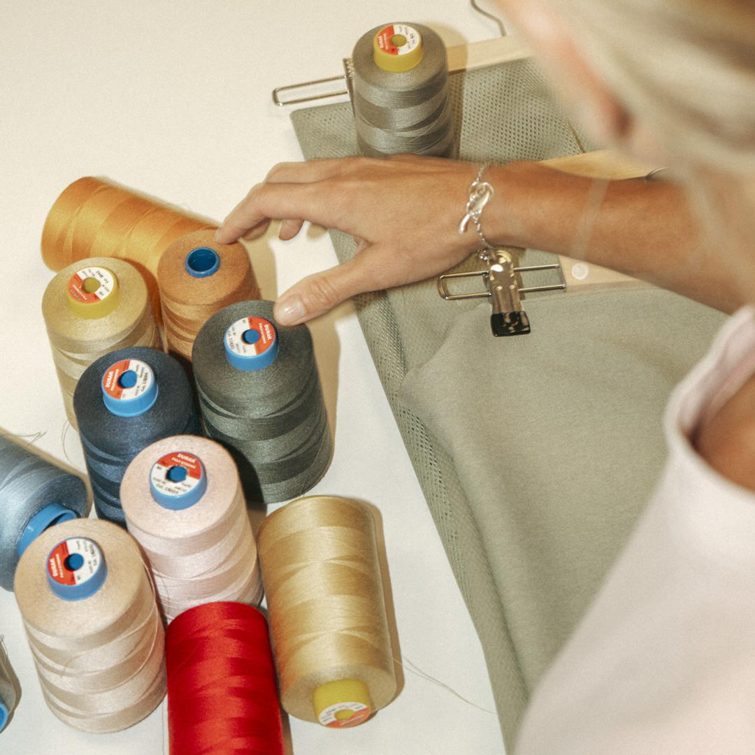 A person selecting thread from a variety of colorful spools on a table, with fabric and a clipboard in the background.