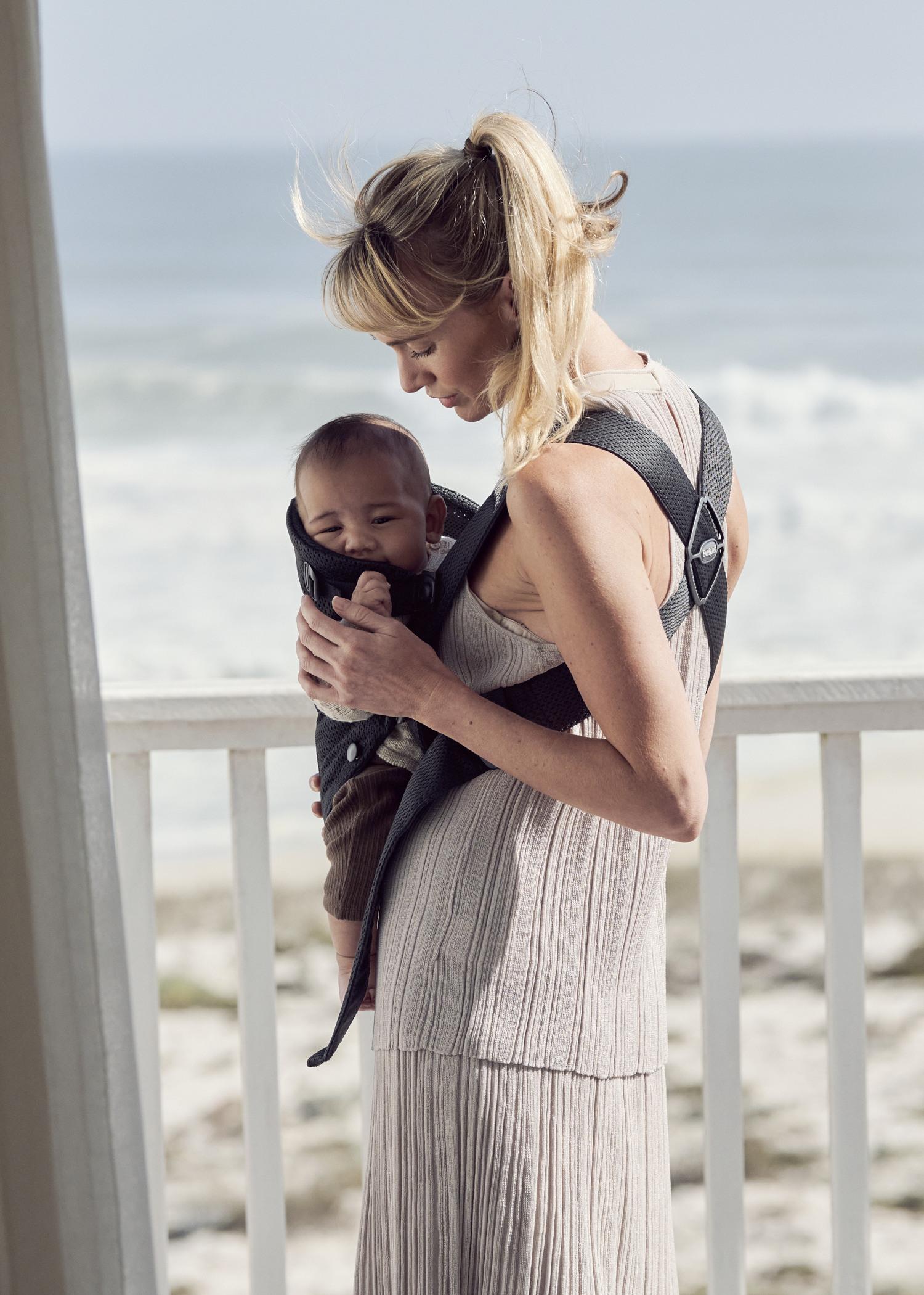 A woman stands on a porch by the ocean, holding a baby in a front carrier. She gazes down lovingly at the child.