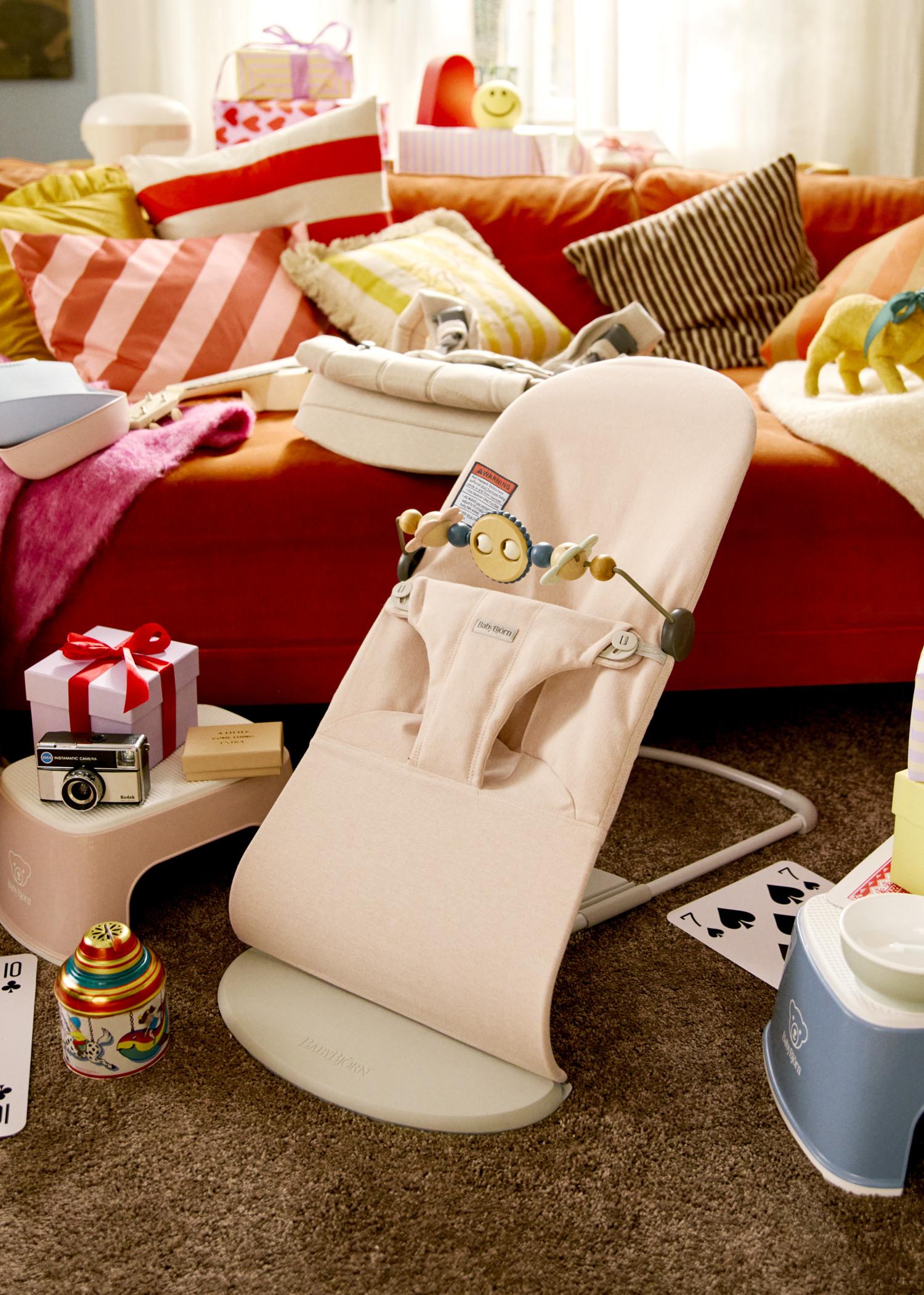 A cozy living room with a pink baby bouncer, toys, a camera, and gifts on a brown carpet. A red sofa with colorful pillows is in the background.