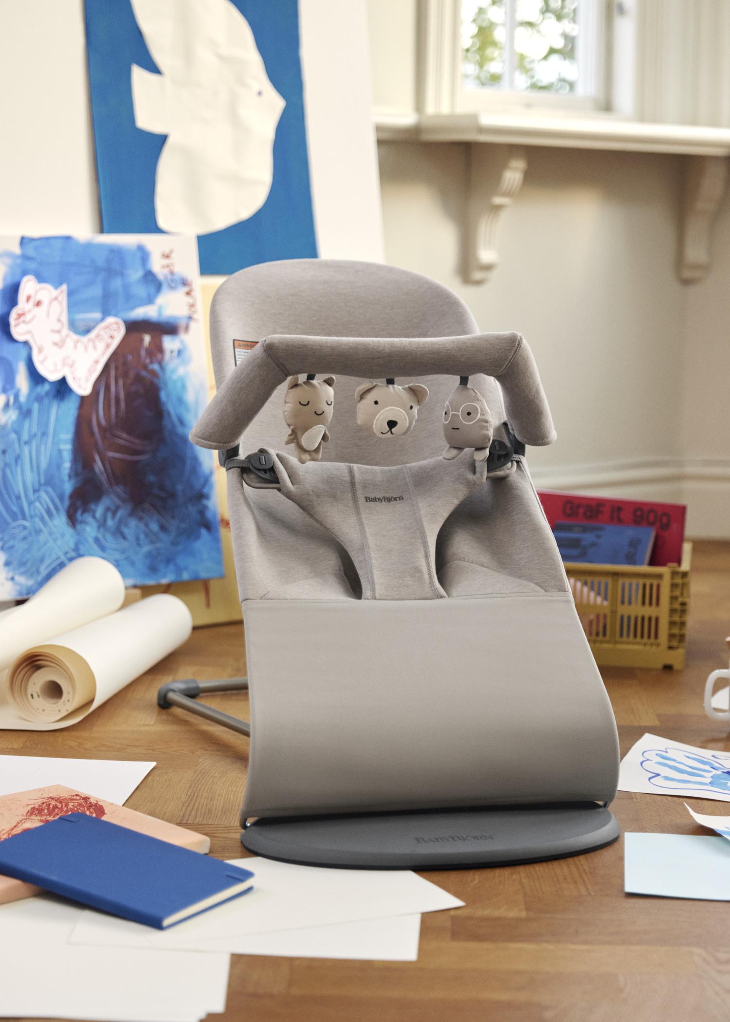 Gray baby bouncer with toy bar featuring animal faces, surrounded by colorful children's drawings and art supplies on a wooden floor.