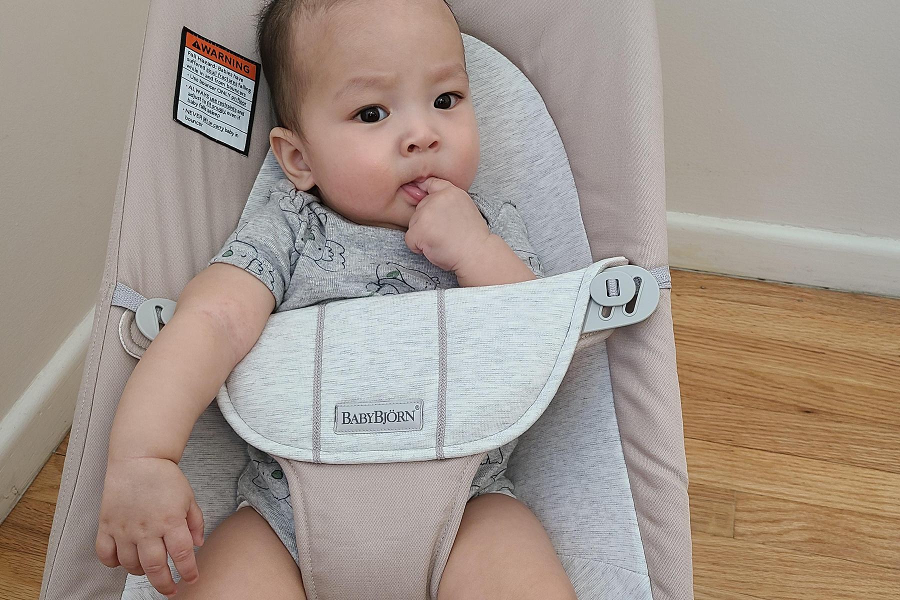 Baby in a gray onesie sits in a beige baby bouncer, sucking thumb, with a curious expression. Wooden floor and light-colored wall in the background.
