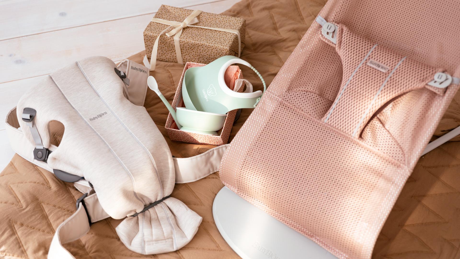 Baby items on a quilted mat: a beige carrier, pink bouncer, pastel feeding set, and a wrapped gift.