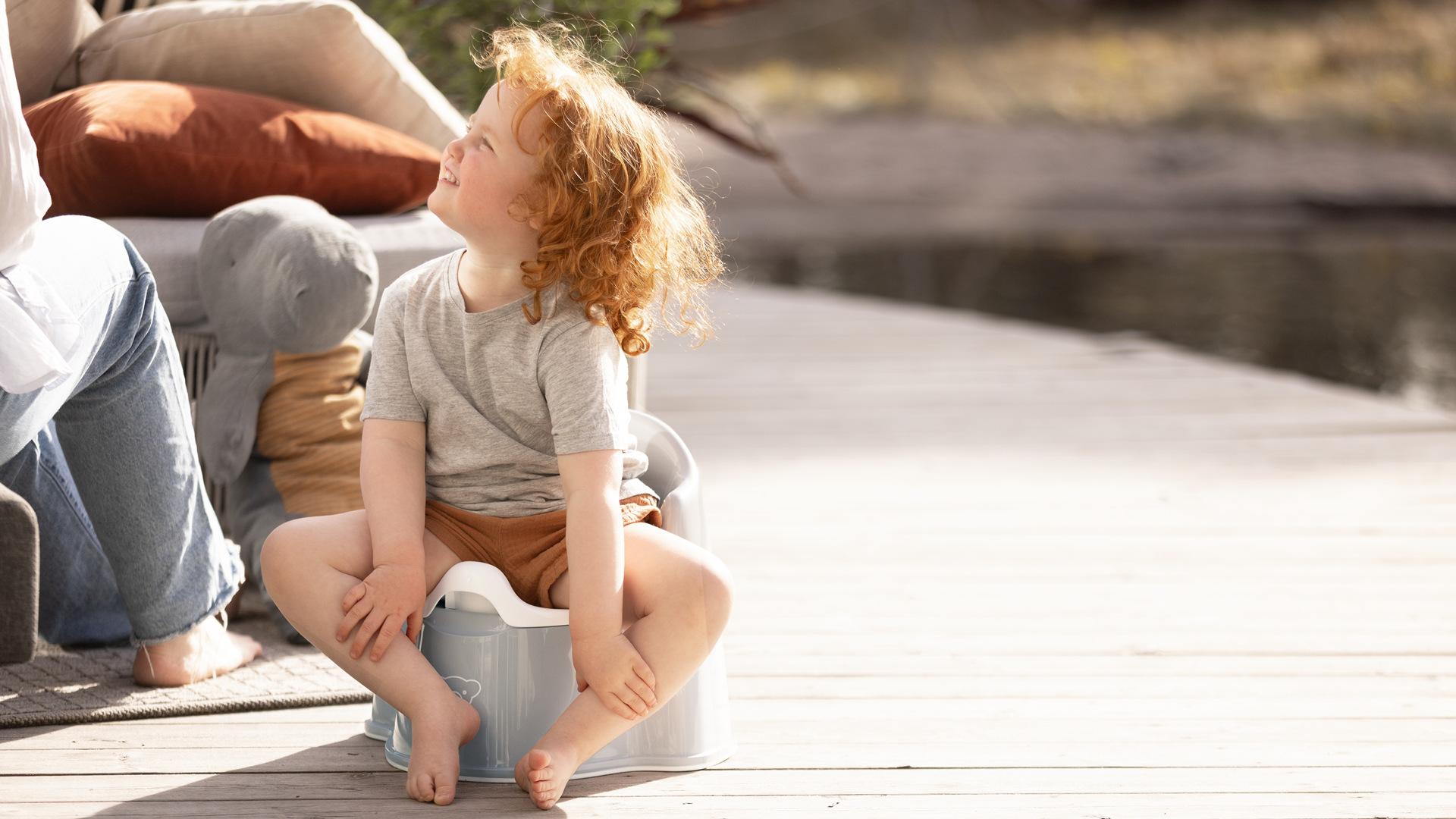 A child with curly red hair sits on a potty outdoors, looking up and smiling. The setting is a wooden deck with pillows and soft lighting.