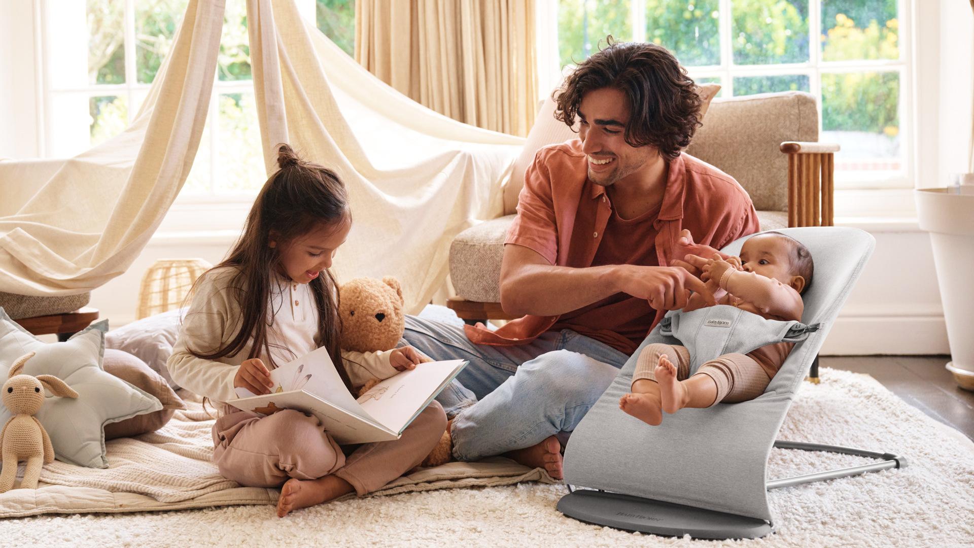 A man sits on the floor with a baby in a bouncer and a young girl reading a book. They are in a cozy, sunlit room with soft furnishings.